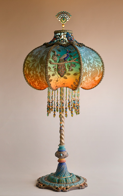 Antique metal table lamp with scroll and leaf motifs has been hand painted and holds a hand-dyed Jeweled Plume Peacock silk lampshade. The shade is dyed from orange into green and blue and covered with antique textiles
